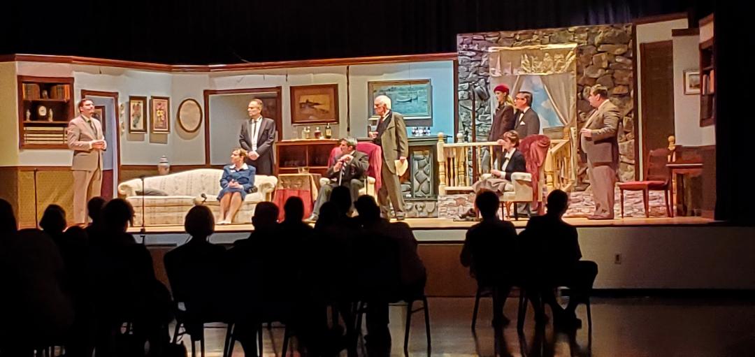 A man in a light brown suit, a woman in a blue skirt sitting on a couch, a man in a light gray suit standing behind her, a man
       in a dark gray suit sitting in a chair, a man in a grayish-green suit standing behind him, a woman in a red cap, a woman in a dark
        jacket sitting in a chair, a man in a dark gray suit, and a man in a brown suit