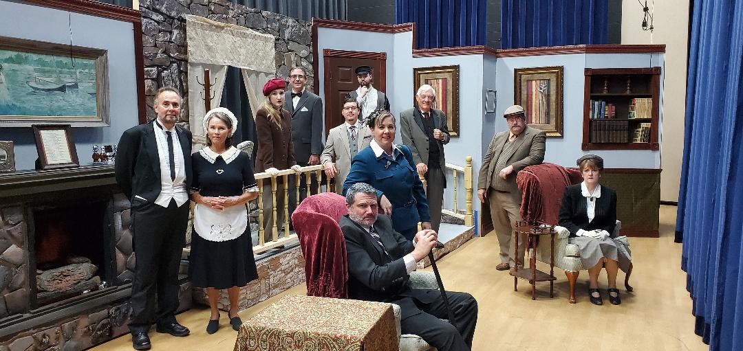 A butler and a maid standing next to a fireplace, a woman in a red cap, a man in a dark gray suit, all standing; a man in a dark gray suit
       sitting in a chair, a woman in a blue dress behind hime, a man in a light gray suit behind her, a man in a graying-green suit
       next to him, a man in a black cap behind both of them; a man in a brown suit, and a woman in a black jacket sitting in a chair