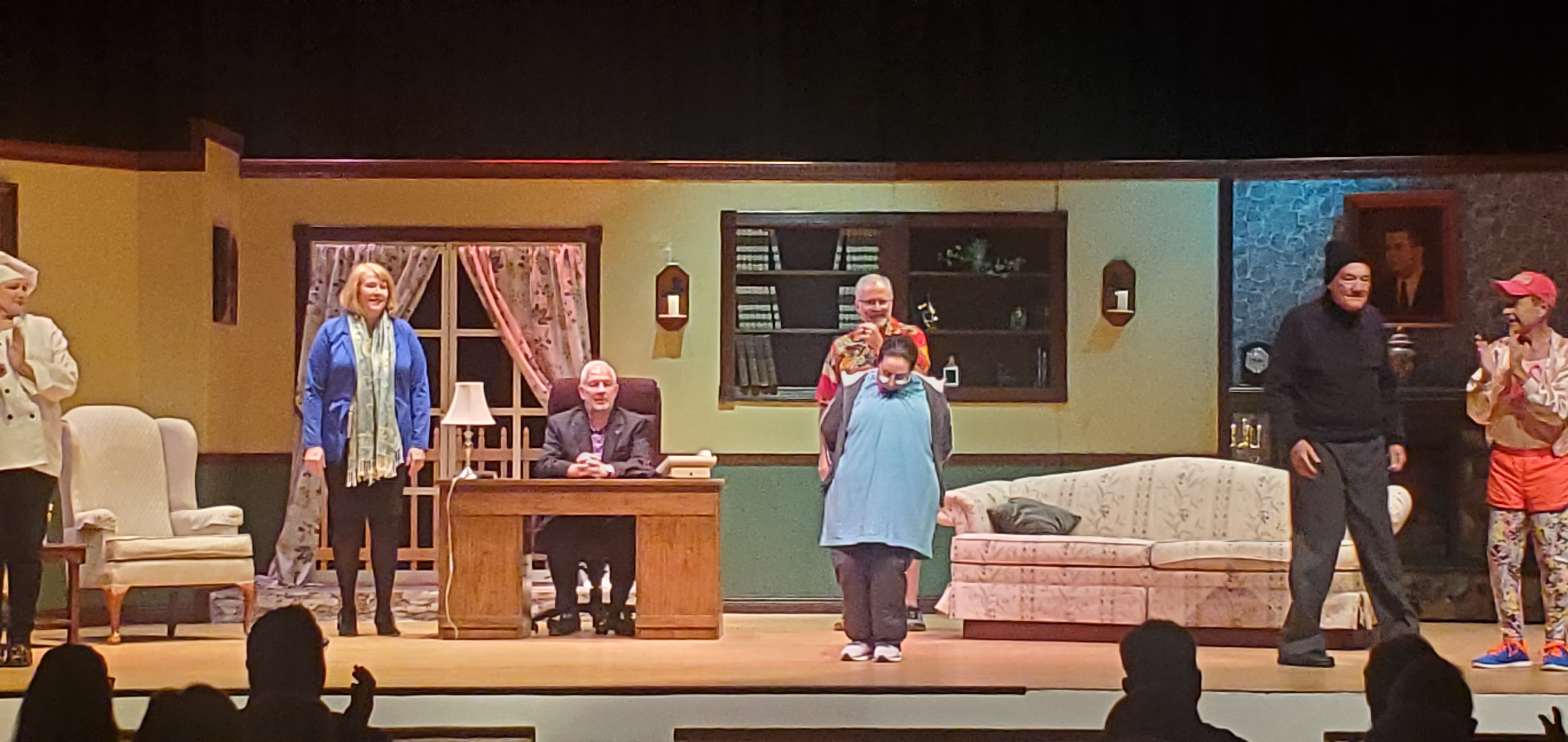 From left to right: a chef in a tope, a woman in a blue jacket and light green scarf, a man in a black suit sitting behind a desk, a woman in a long bluish green top standing in front of a man in a red Hawaiian shirt and brown shorts,
       a man in a black pullover, gray pants and black cap, and a man in an orange running outfit clapping.