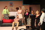 A man sitting on a couch, a woman wearing a green sweater, and a woman in a white dress, two women in black pantsuits,
       a woman in a black dress, and a man wearing a hat and a tie