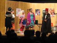 A woman in a black dress and mink stole, a man in a sparkling gold shirt, a woman in a red, white and blue blouse and dress, a woman in a
       sparkling purple coat, and a man in a suit, black cloak and red turban