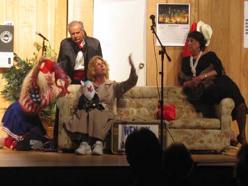 A woman in a red, white and blue dress kneeling next to a couch on which a woman in a brown raincoat and a woman in a black
       dress sit, with a man in a suit standing behind the couch 