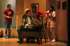 A man and a woman sit at a desk, while a man in a red Avengers T-shirt and a man in a suit stand behind them,
       and a young woman hits a man in a Tampa Bay Buccaneers T-shirt