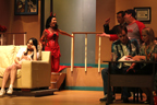A young woman sits on a couch, while a woman dressed like a devil talks to a man in a suit and a man in a Tampa Bay Buccaneers T-shirt, and
       a man a woman sit at a desk