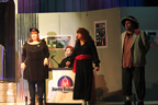 A woman in a black sparkly skirt, a sitting woman dressed in plaid, a man in a black dress and red sash, and a man in a jacket, shirt
       and large hat