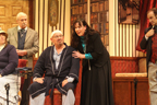 A woman in a dark dress and blue blouse stands behind a bald man with a bandage on his head, while
       Sherlock Holmes looks on from the right, and Dr. Watson and a woman in gray vertical striped dress look on from the left