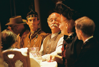 A man in a cowboy hat, a Native American man, a bald man, a woman in a hat, a man in a suit, and a woman with her back
       to the camera all sitting a table
