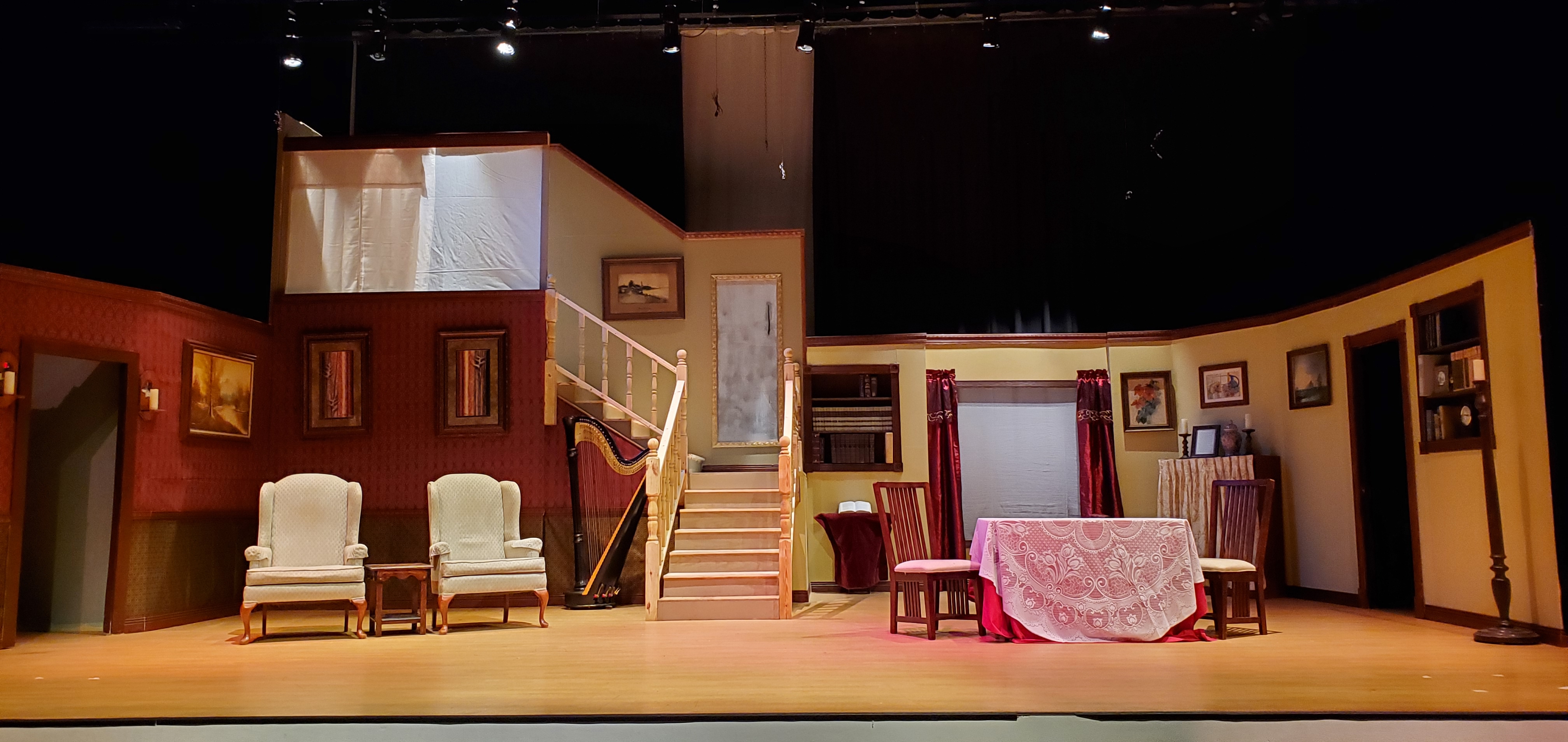 A theater set with reddish brown walls on the left and light brown walls on the right, and a set of stairs just to the left of center. The stairs turn
       left at a landing and go up again. A harp sits near the stairs. Two beige chairs sit to the left and a table and two chairs sit toward the right. There is a
       large window with red drapes toward the back on the right. There are doorways to the left and right.