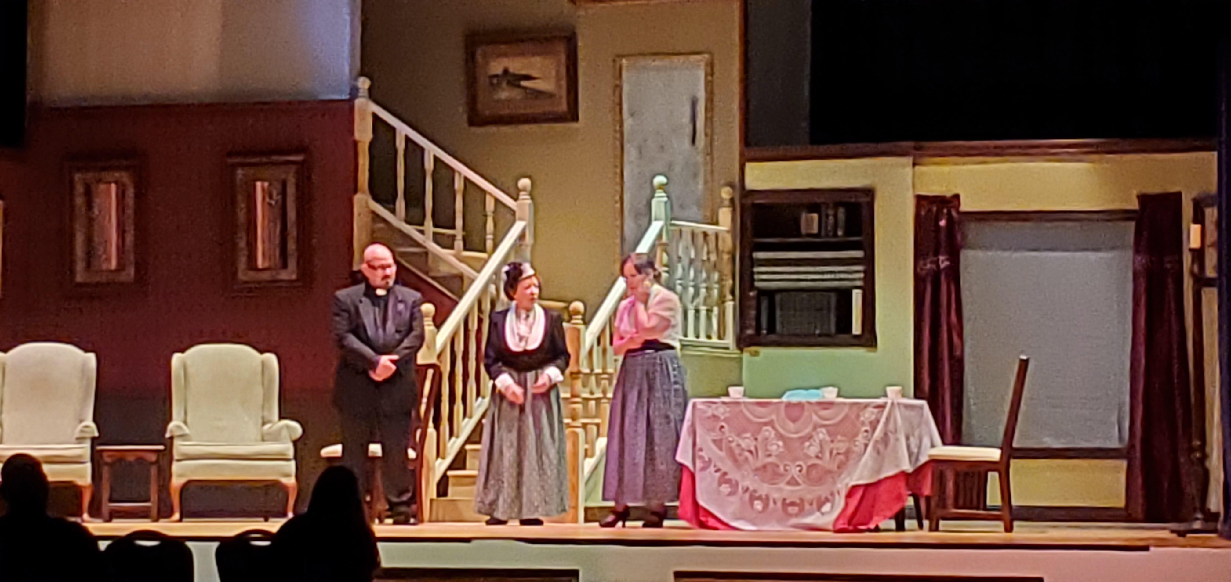A priest, a woman in Victorian dress and another woman in a white blouse and a long gray dress stand near the steps. A table with a white tablecloth
       and and rose-colored cloth underneath is to the right of the people.