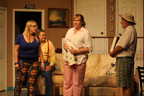 A woman wearing brightly-colored stretch pants, a man in a yellow shirt and red suspenders sitting on a couch, a woman in pink pants, and a man in plaid shorts